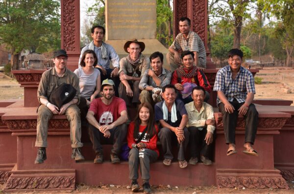 Team of archaeologists in the field sitting for a group photo in front of pillar monument