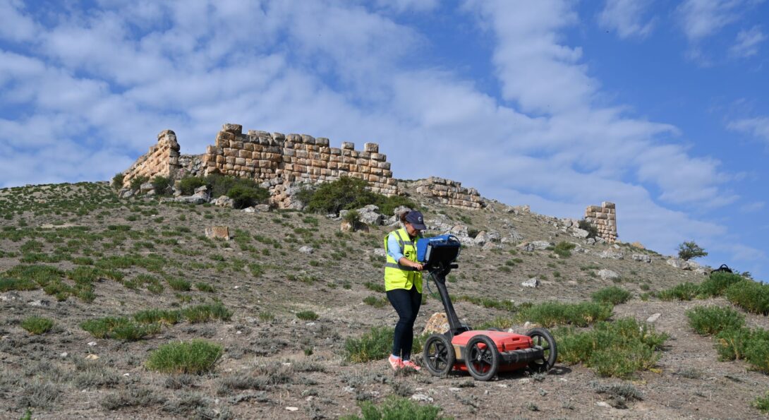 Archaeologist conducting geophysical survey on a low hill with stone ruins at the top on a bright sunny day