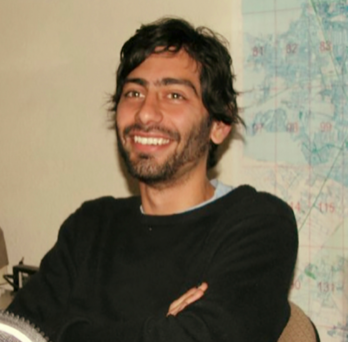 Dr. Solinis Casparius (man with facial hair, dark hair, in a black sweater, with arms crossed) in front of a map
                  