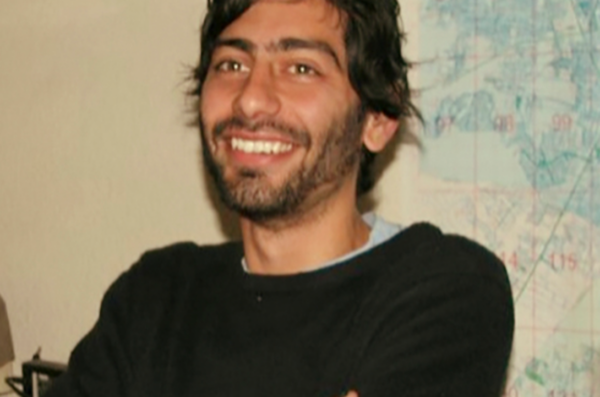 Dr. Solinis Casparius (man with facial hair, dark hair, in a black sweater, with arms crossed) in front of a map